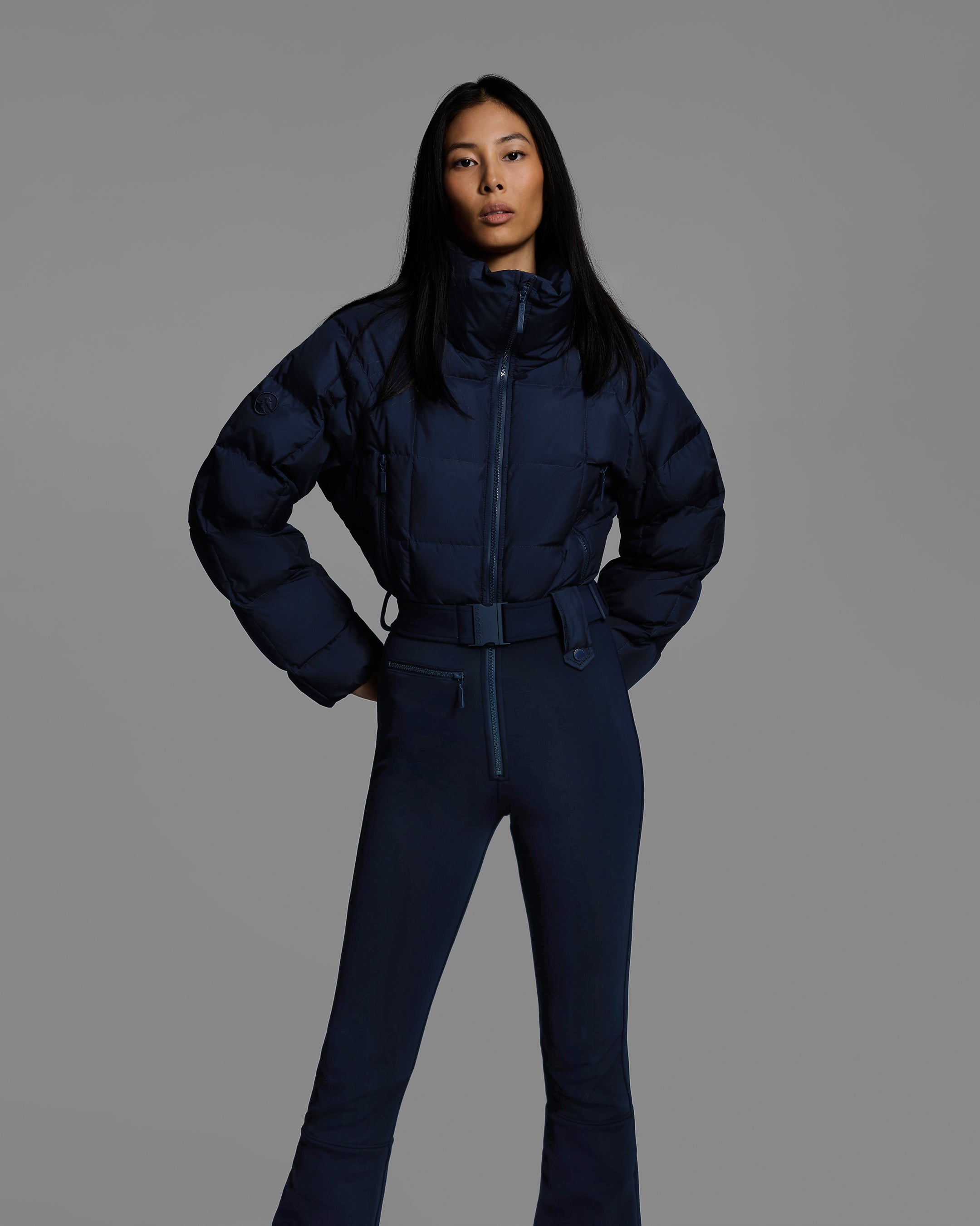 Front detail view of a female model wearing a belted indigo blue color ski suit with quilted, down filled top and 4-way stretch bottom. #color_indigo