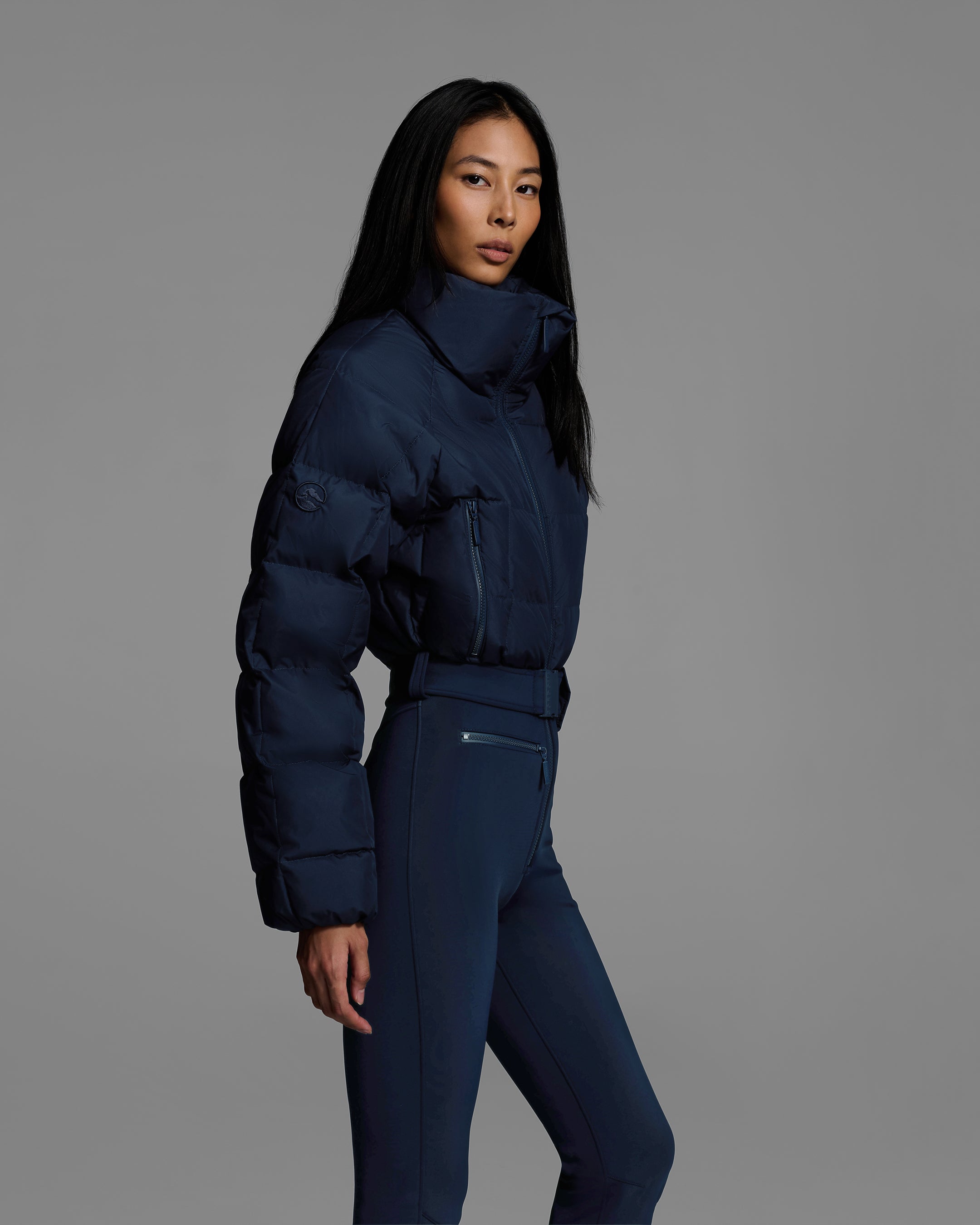 Side detail view of a female model wearing a belted indigo blue color ski suit with quilted, down filled top and 4-way stretch bottom. #color_indigo