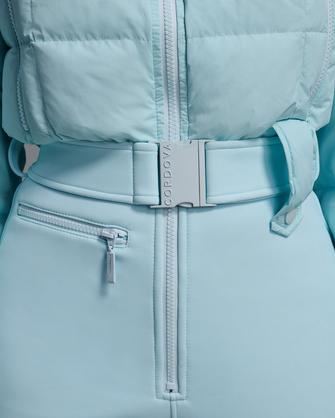 Close up detail view of an iceberg blue ski suit with a quilted down top, belted waist, and 4-way stretch pant. #color_iceberg