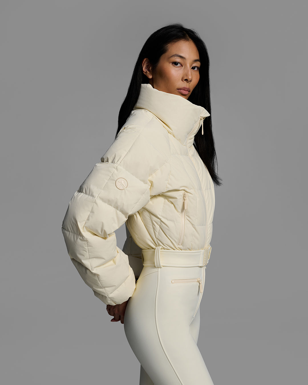Side detail view of a female model wearing a belted alabaster white color ski suit with quilted, down filled top and 4-way stretch bottom. #color_alabaster