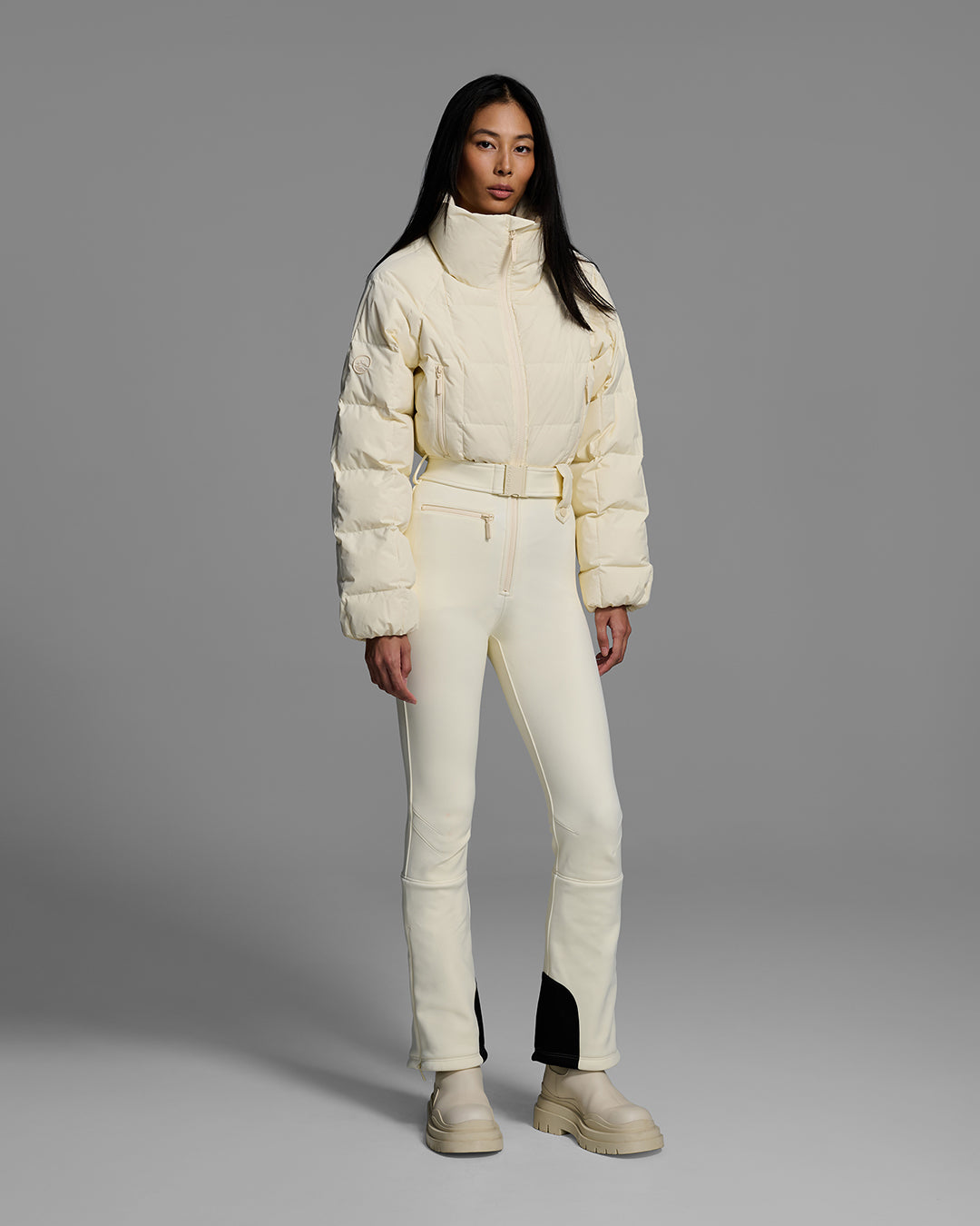 Female model wearing a belted alabaster white color ski suit with quilted, down filled top and 4-way stretch bottom. #color_alabaster