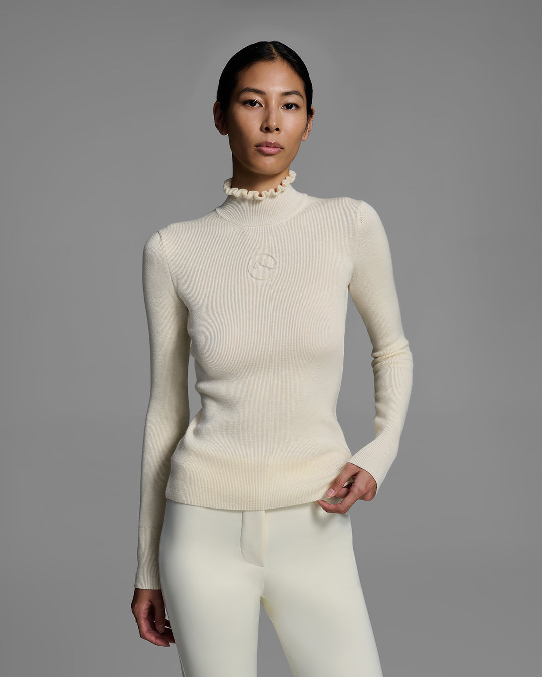 Woman wearing a white ski sweater with a high ruffle neck and 3D mountain logo on chest. #color_alabaster
