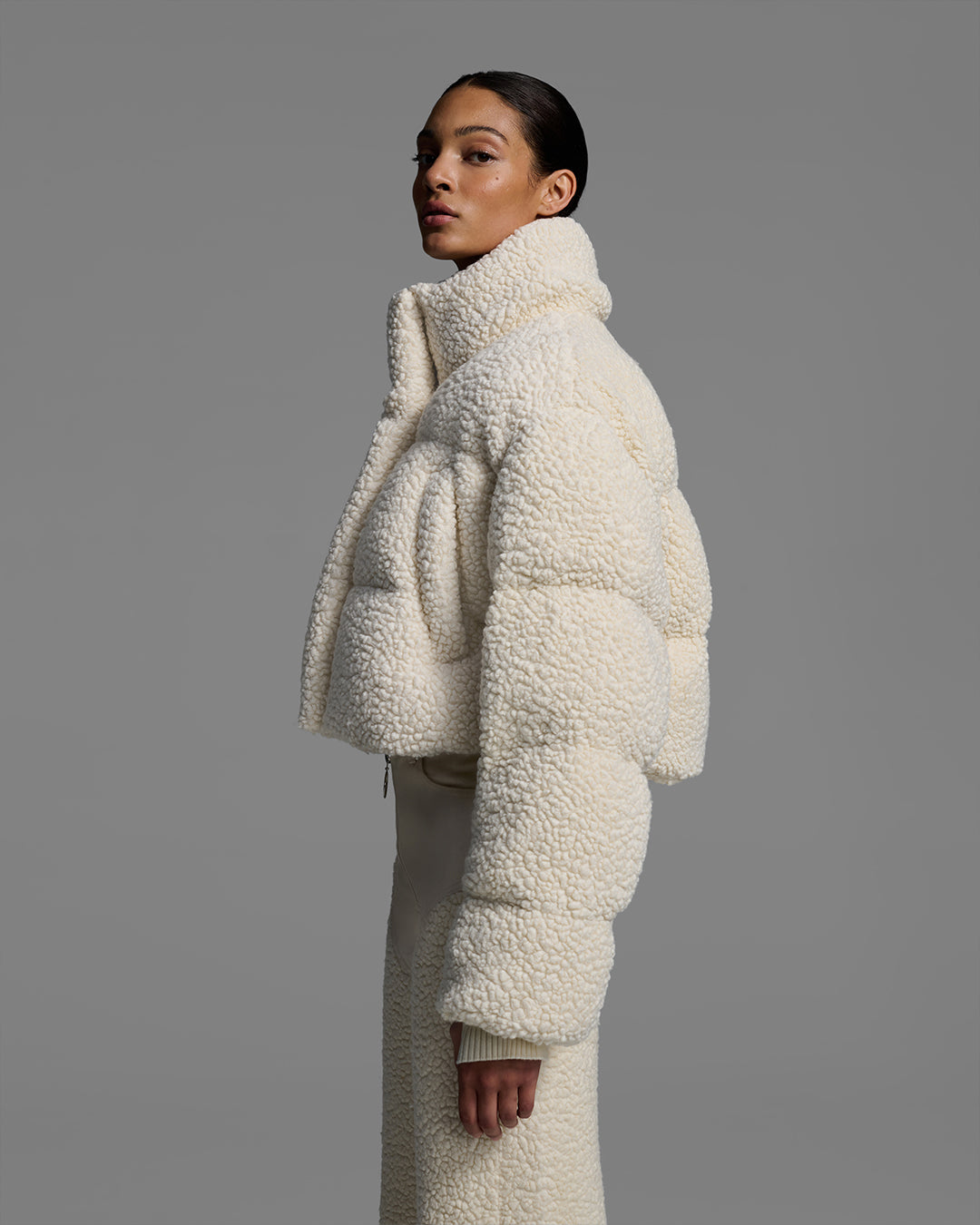 Side view of female model wearing a natural, off-white, sherpa puffer with matching pant on a gray background. #color_natural