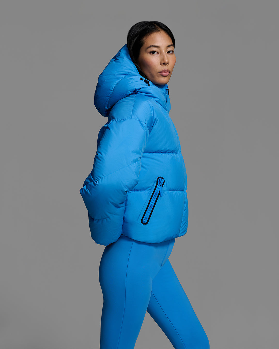 Side detail view of a female model wearing a classic down filled ski jacket in bright blue with a matching ski pant. #color_azul