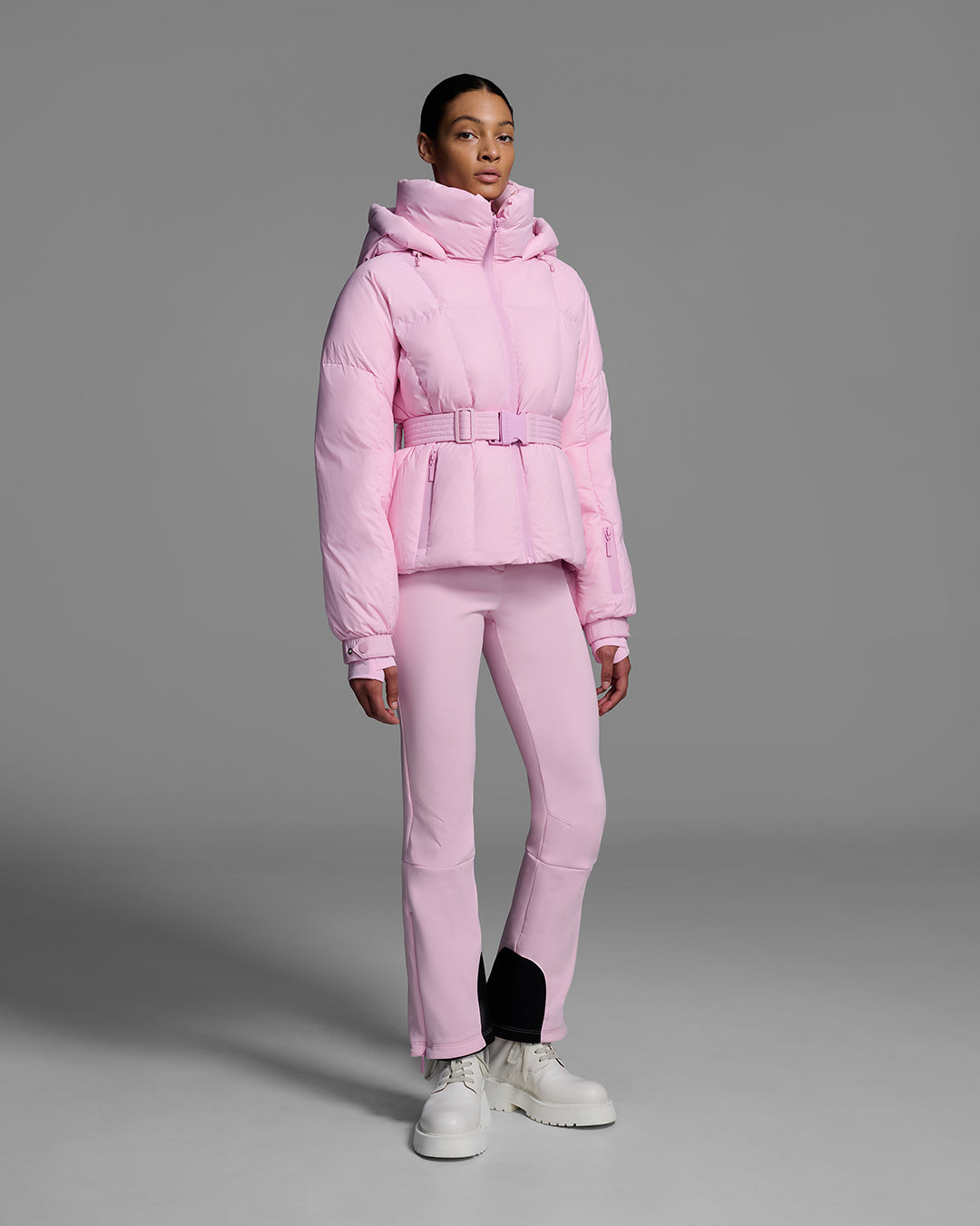 Female model wearing a classic slope style down filled, belted ski jacket in peony pink color with a matching ski pant. #color_peony