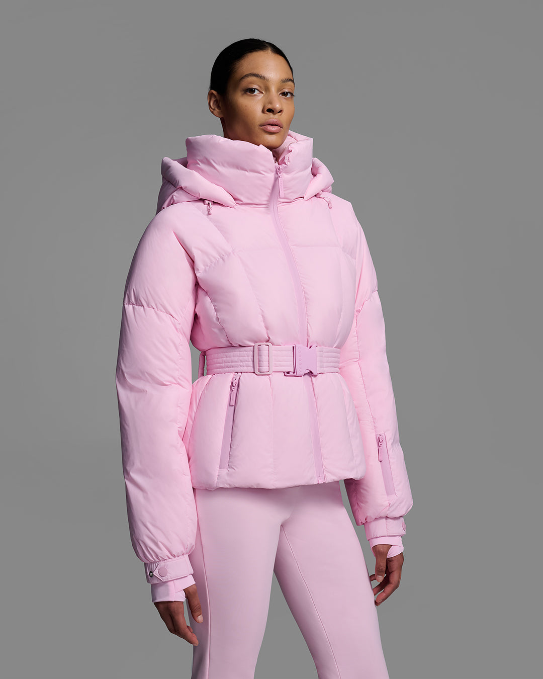 Detail front view of a female model wearing a classic slope style down filled, belted ski jacket in peony pink color with a matching ski pant. #color_peony