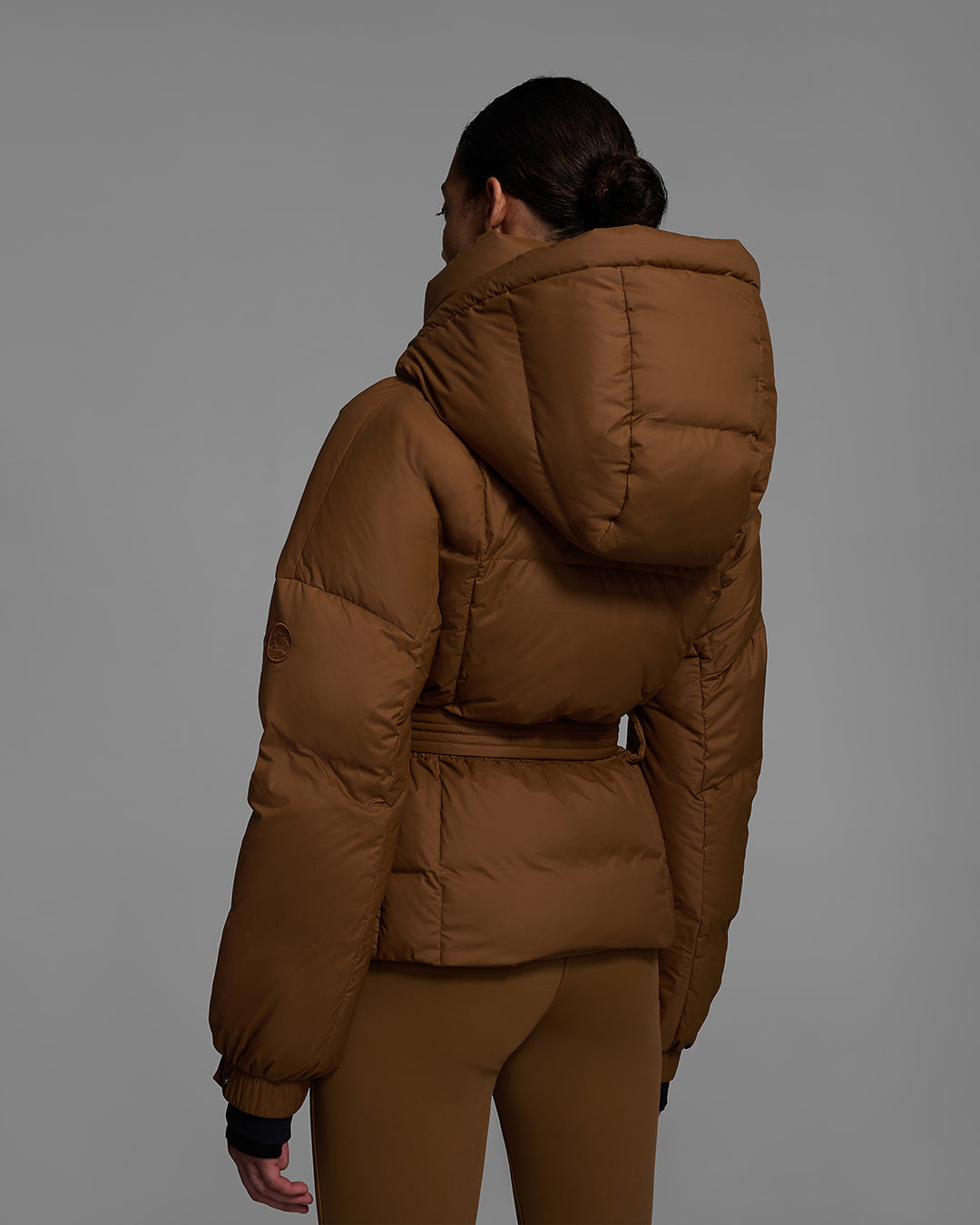 Back view of a female model wearing a classic slope style down filled, belted ski jacket in sepia brown color with a matching ski pant. #color_sepia
