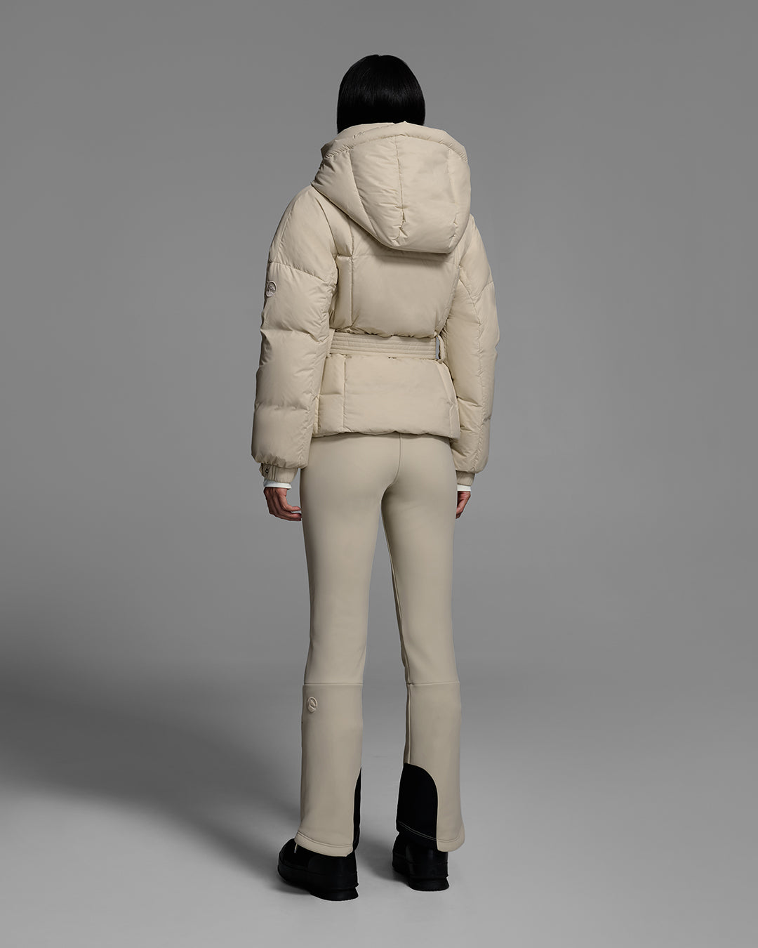 Back view of a female model wearing a classic slope style down filled, belted ski jacket in birch inspired tan color with a matching ski pant. #color_birch