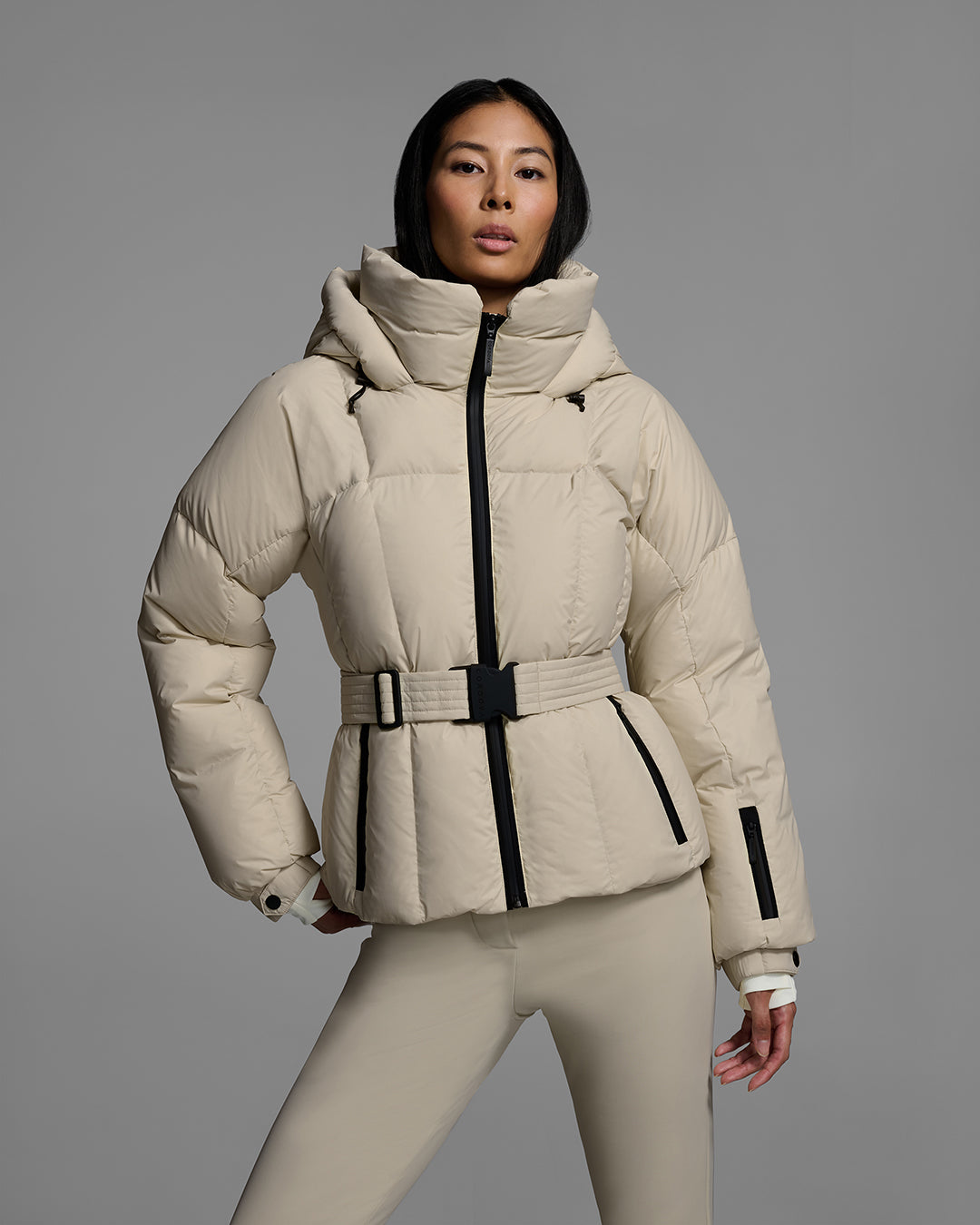 Detail front view of a female model wearing a classic slope style down filled, belted ski jacket in birch inspired tan color with a matching ski pant. #color_birch