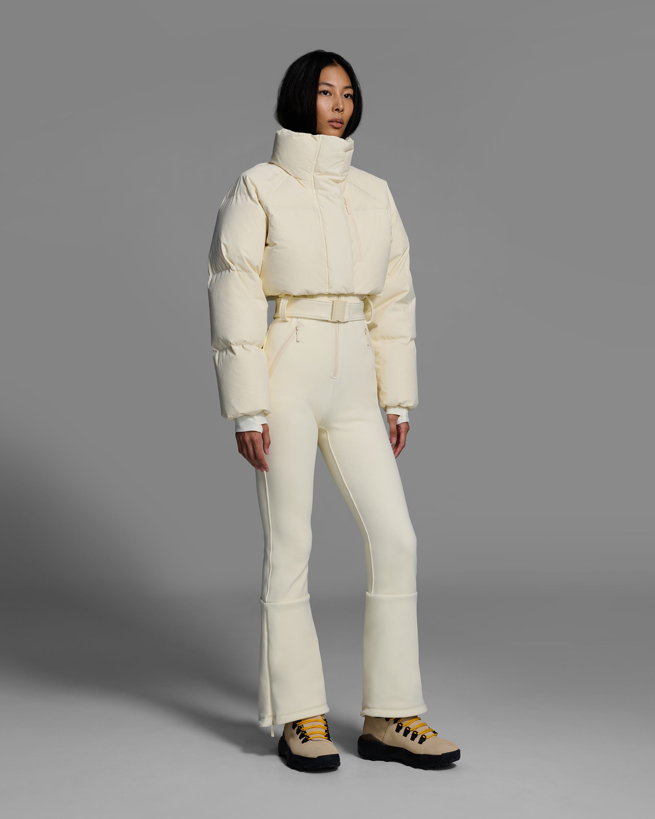 Female model wearing an alabaster white ski suit with a white puffer crop top, belted waist, and 4-way stretch pant. #color_alabaster