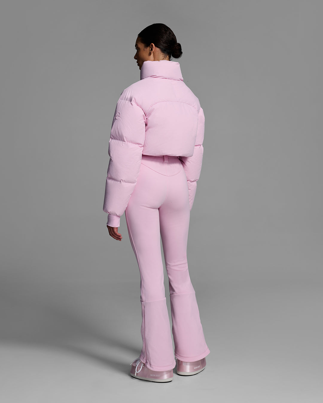 Back view of a female model wearing a peony pink ski suit with a puffer crop top, belted waist, and 4-way stretch pant. #color_peony