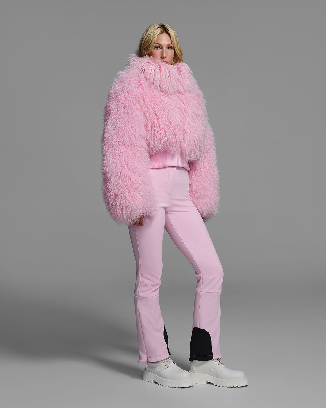 Female model wearing a natural shearling jacket in peony pink with matching ski pant. #color_peony
