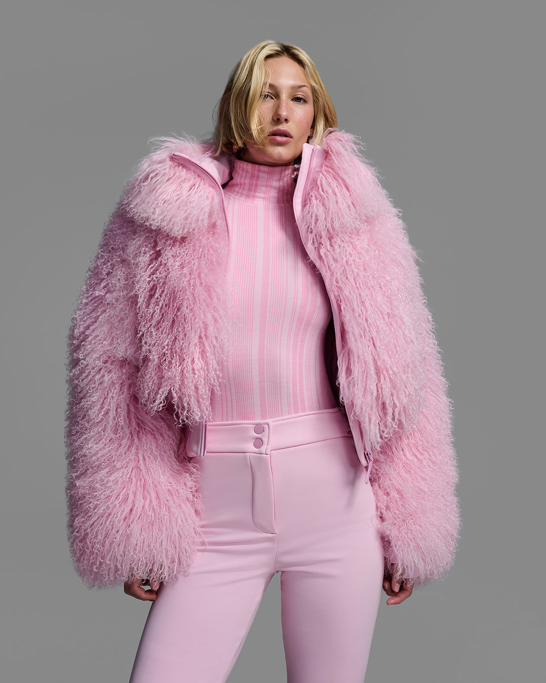 Female model wearing a natural shearling jacket, unzipped, in peony pink with matching ski pant and base layer top. #color_peony
