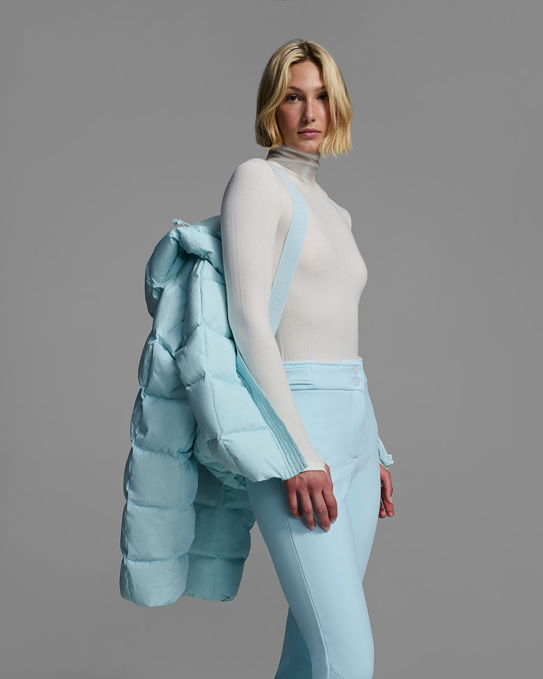 Woman wearing a iceberg blue color quilted down ski jacket like a backpack with it's interior backpack straps, with matching ski pants. #color_iceberg