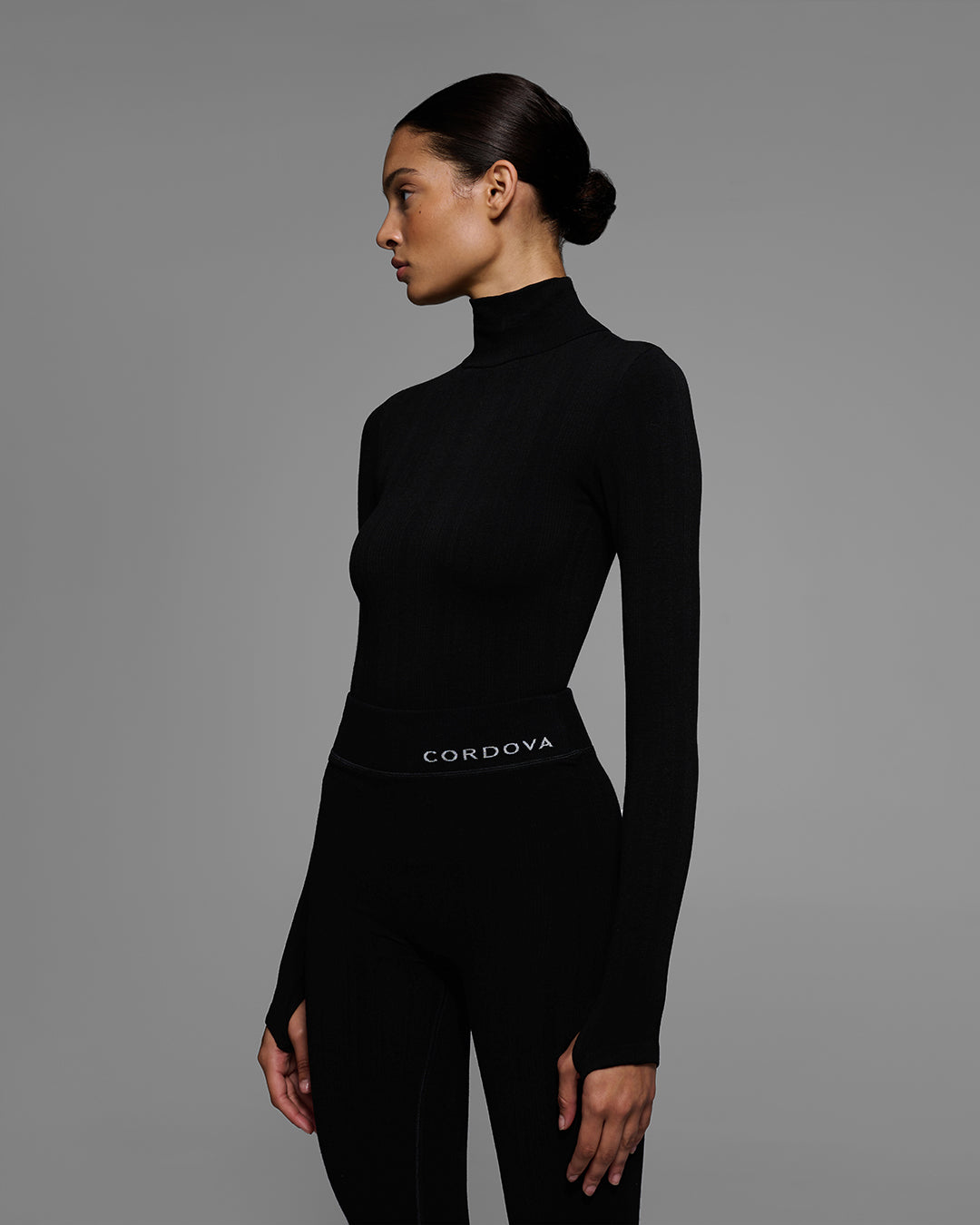 Detail view from the knees up of a woman wearing a black turtleneck and matching pant base layer set made of merino wool blend. #color_onyx