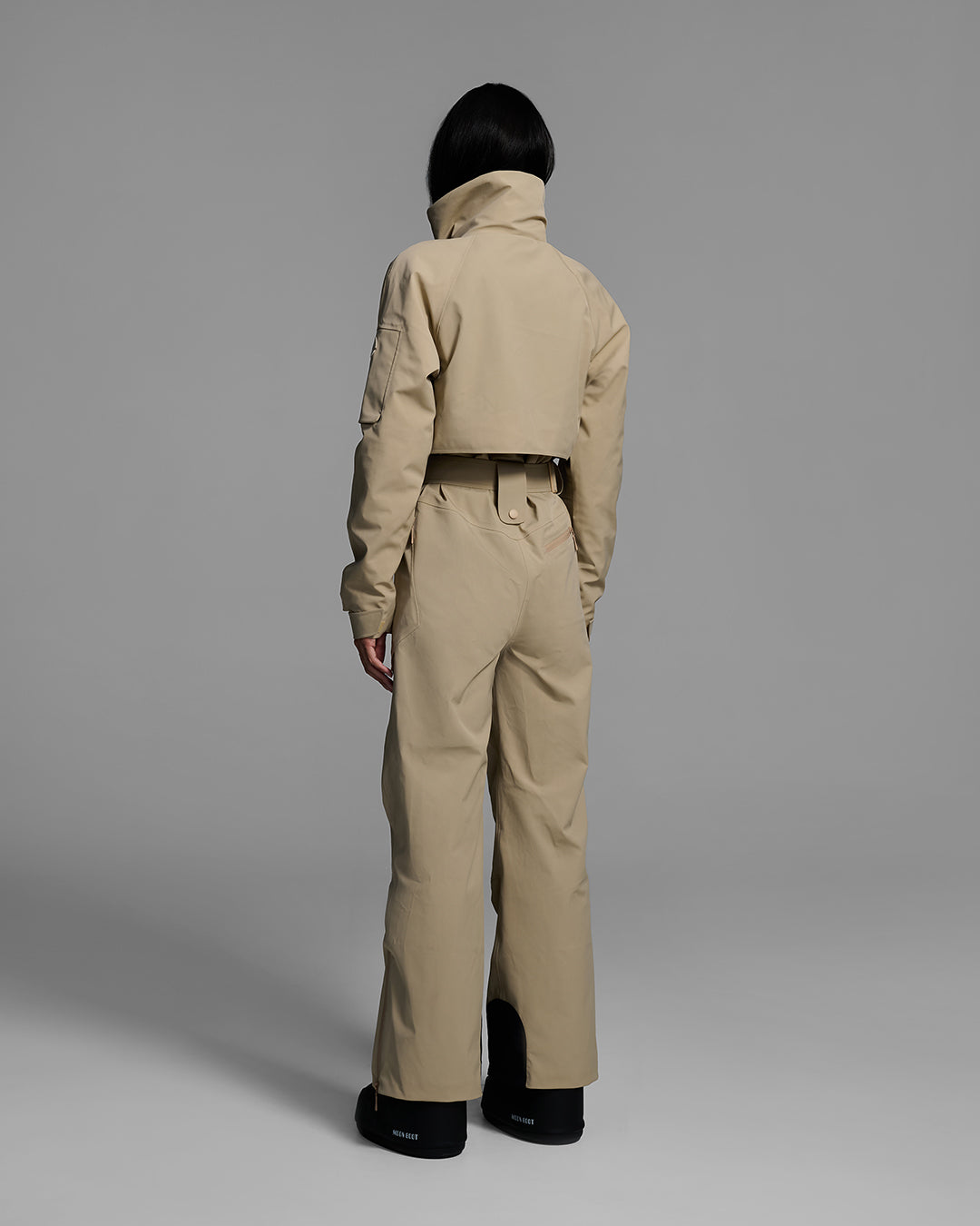 Back view of a woman wearing a tan color waterproof technical fabric ski suit with a belted waist. #color_sand