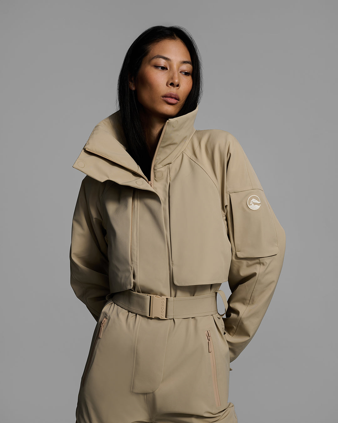 Detail view of a woman wearing a tan color waterproof technical fabric ski suit with a belted waist. #color_sand